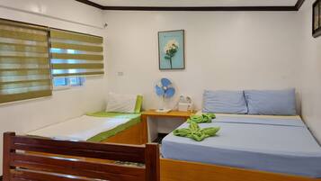 Una habitación pequeña y luminosa con una cama, una silla, una mesita de noche con un ventilador y una ventana con persianas.
