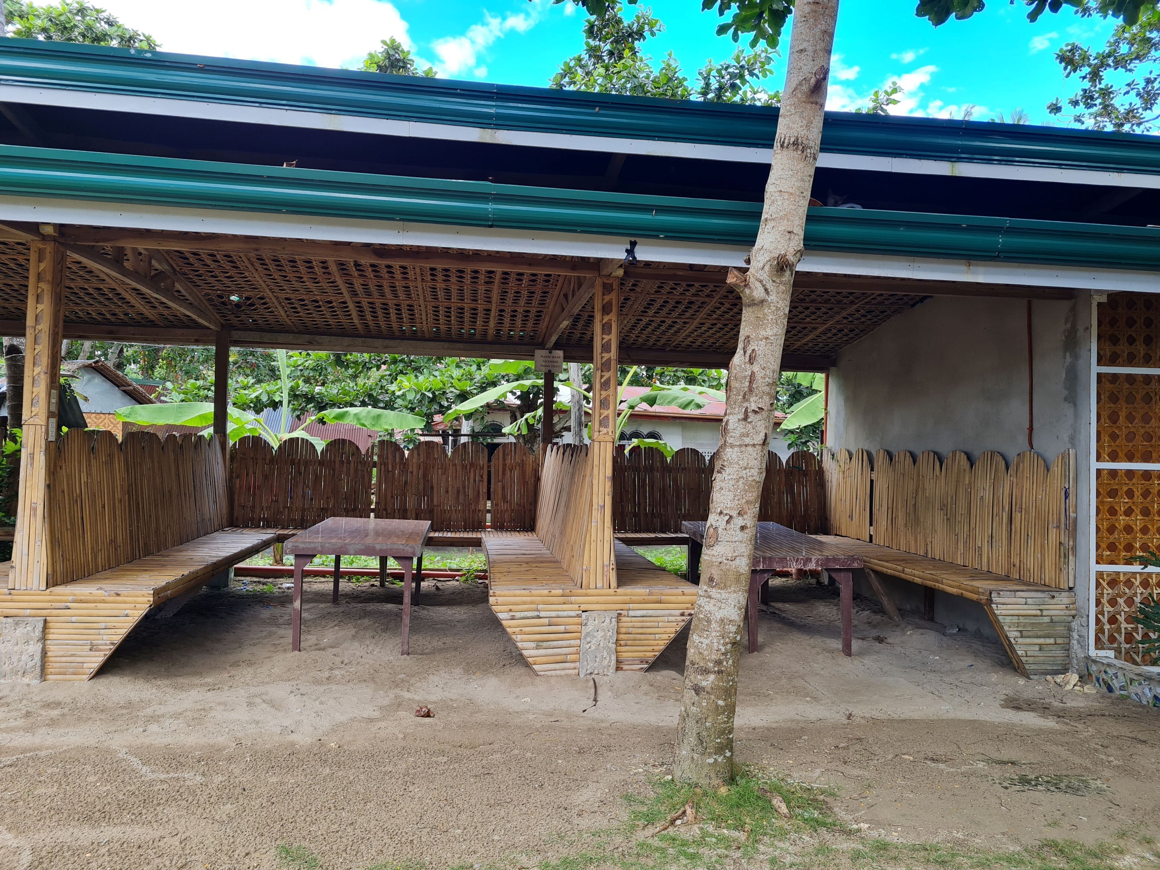 A covered outdoor seating area with benches and tables, surrounded by a wooden fence and a tree.