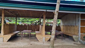 A covered outdoor seating area with benches and tables, surrounded by a wooden fence and a tree.