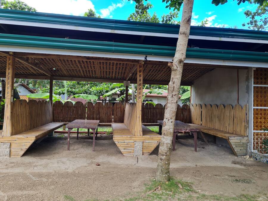 A covered outdoor seating area with benches and tables, surrounded by a wooden fence and a tree.