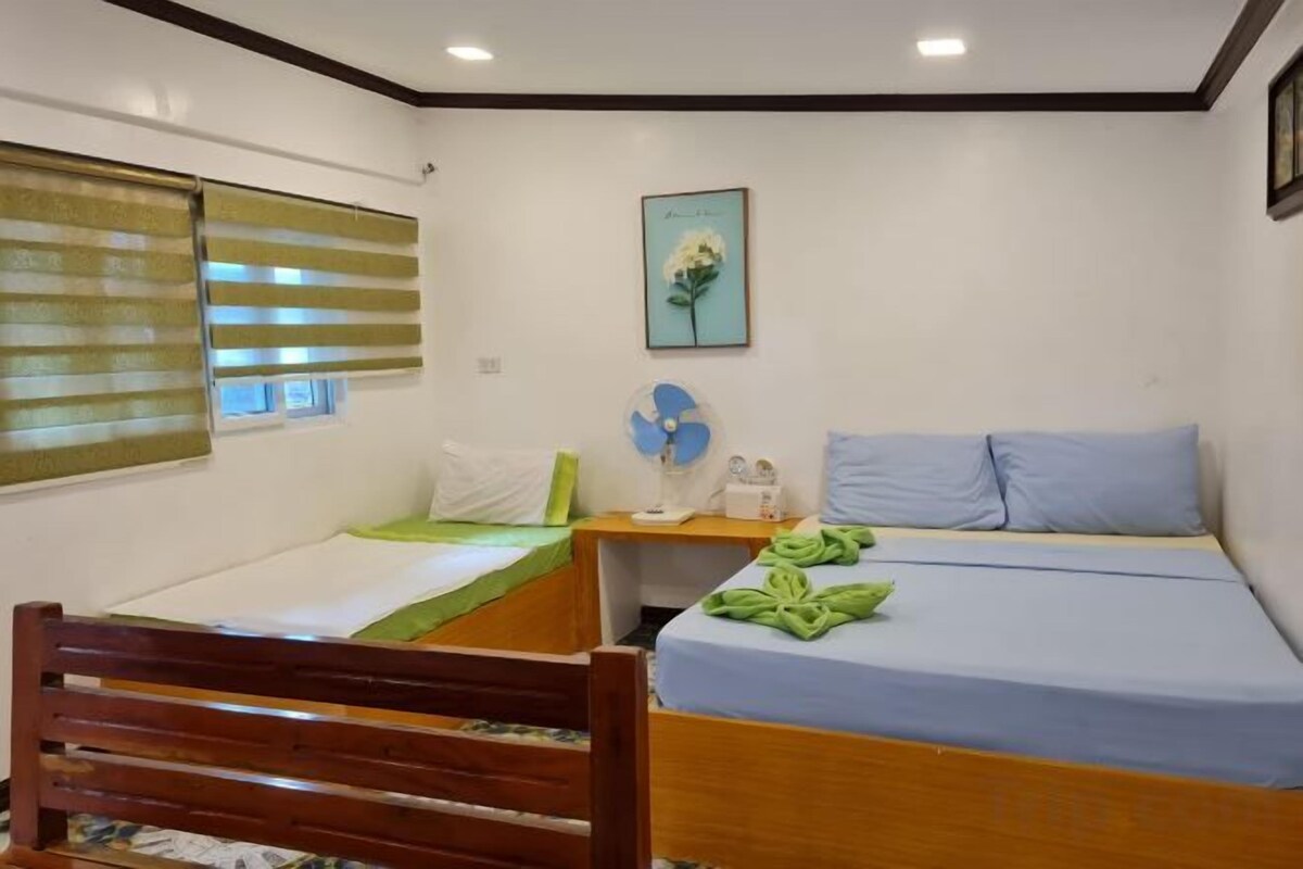 A room with two beds, a wooden chair, a bedside table, a fan, and a picture on the wall.