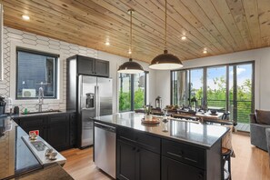 A modern kitchen with a central island, black cabinetry, stainless steel appliances, and large windows offering views of greenery.