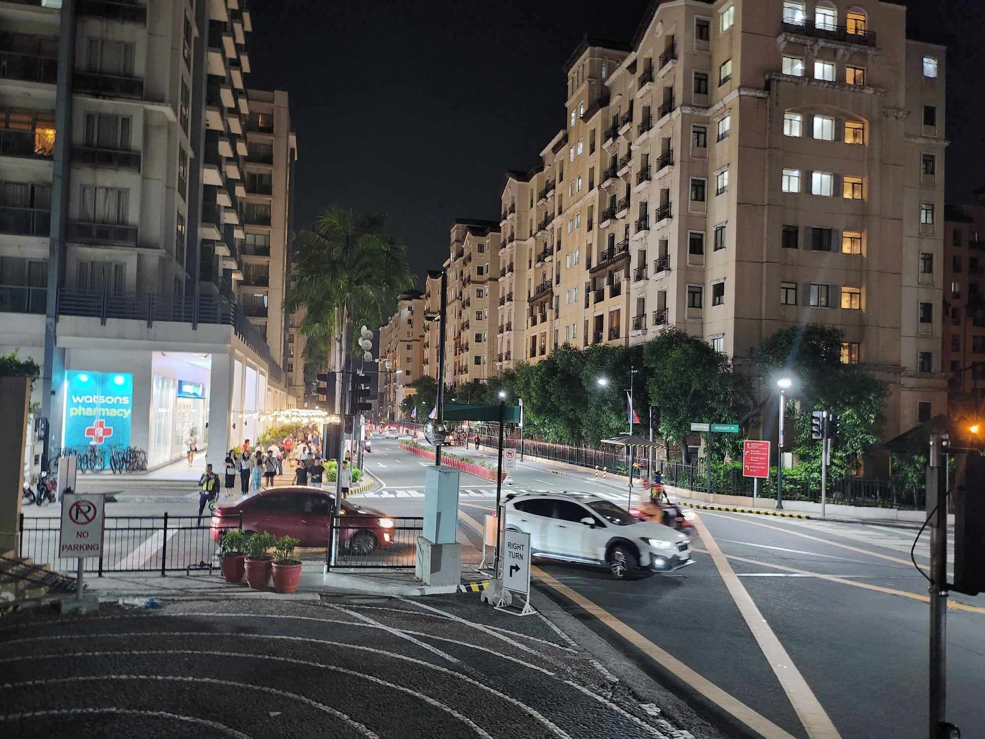 Una calle urbana nocturna con edificios residenciales de varios pisos, una farmacia y un paso de peatones.