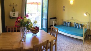 Une pièce avec une table à manger en bois, un panier de fruits, un vase de fleurs, un lit avec des coussins à rayures et une lampe fixée au mur.