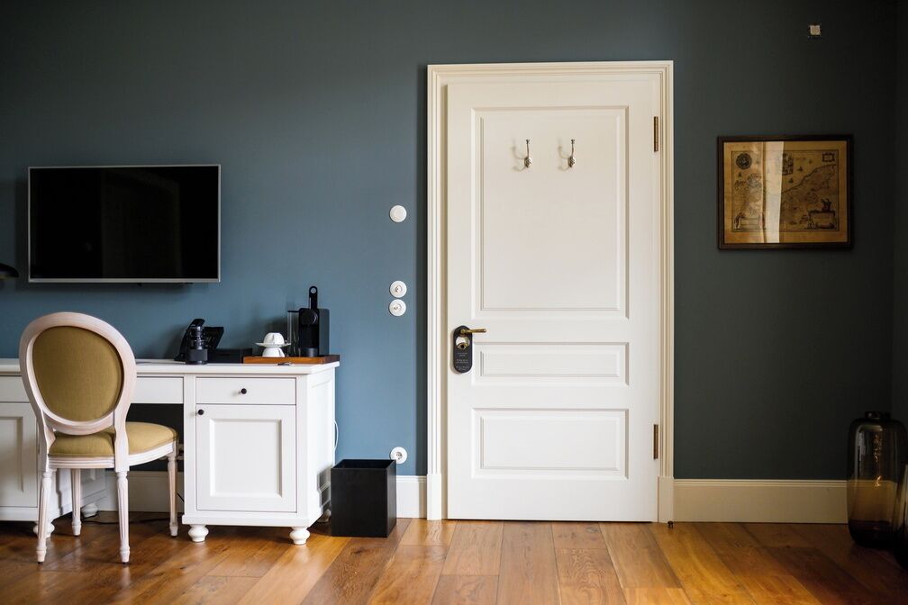 A room with a white desk, a chair, a television mounted on the wall, and a white door with two hooks.