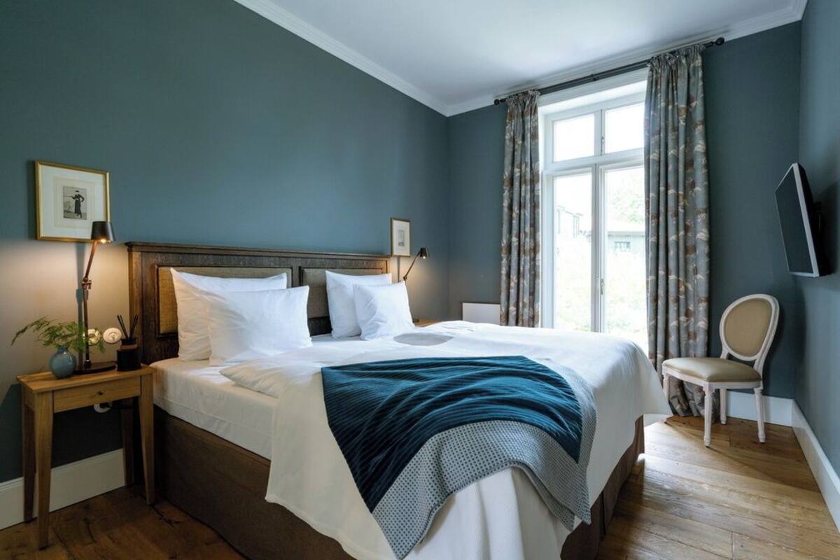 A neatly made bed with white linens, a teal throw blanket, and a wooden headboard. There is a bedside table with a lamp and a small plant. A chair is positioned near the window, which has floral curtains.