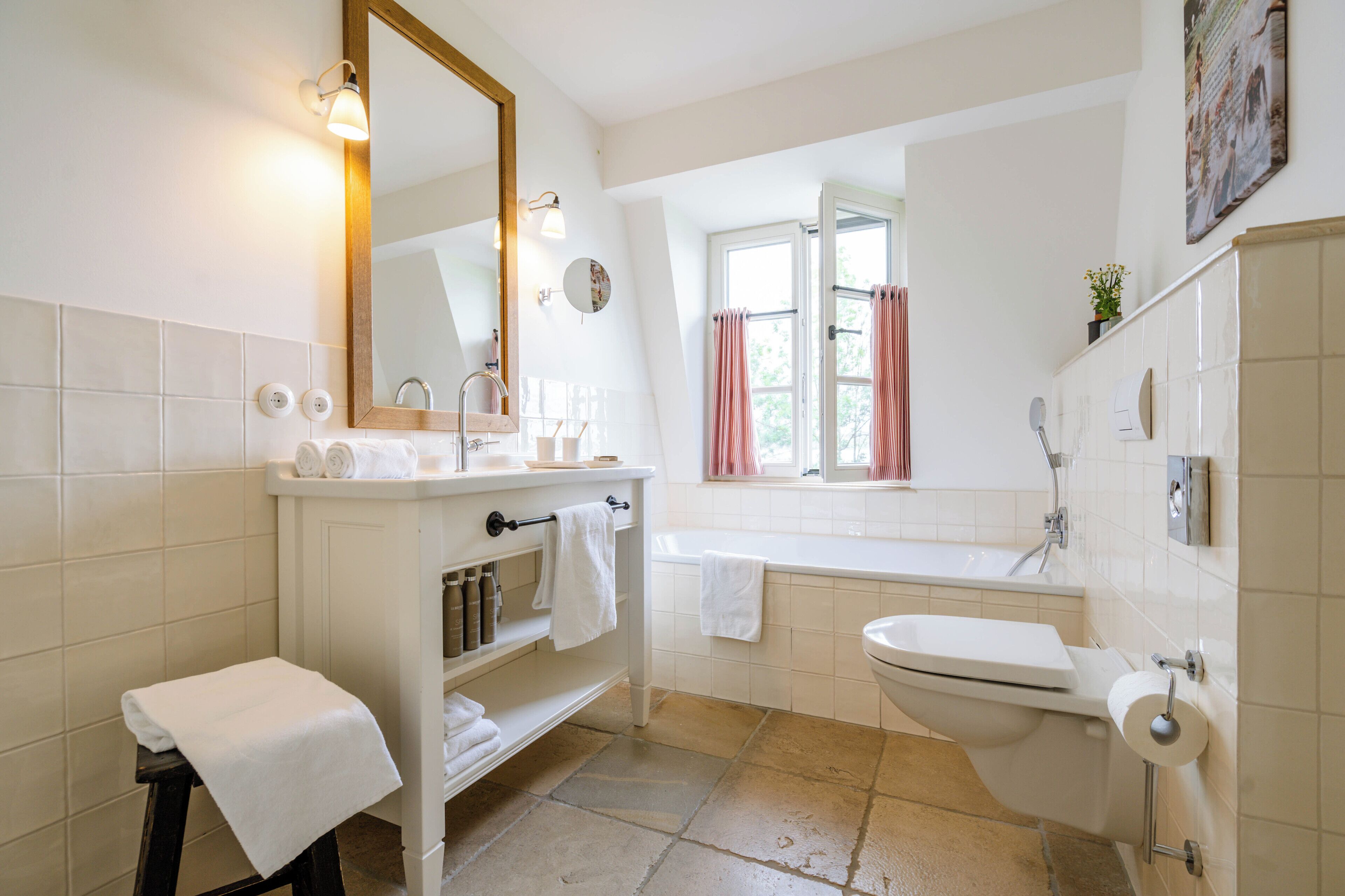 Un baño moderno con lavamanos, inodoro y bañera blancos. Hay un mueble de tocador con un espejo y una repisa con artículos de aseo. Se ve una ventana con cortinas rosadas, y el piso está cubierto de baldosas.