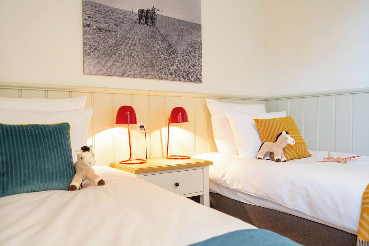 A hotel room with two beds, each with white bedding and a small bedside table with a lamp. There is a wall-mounted picture above the beds.