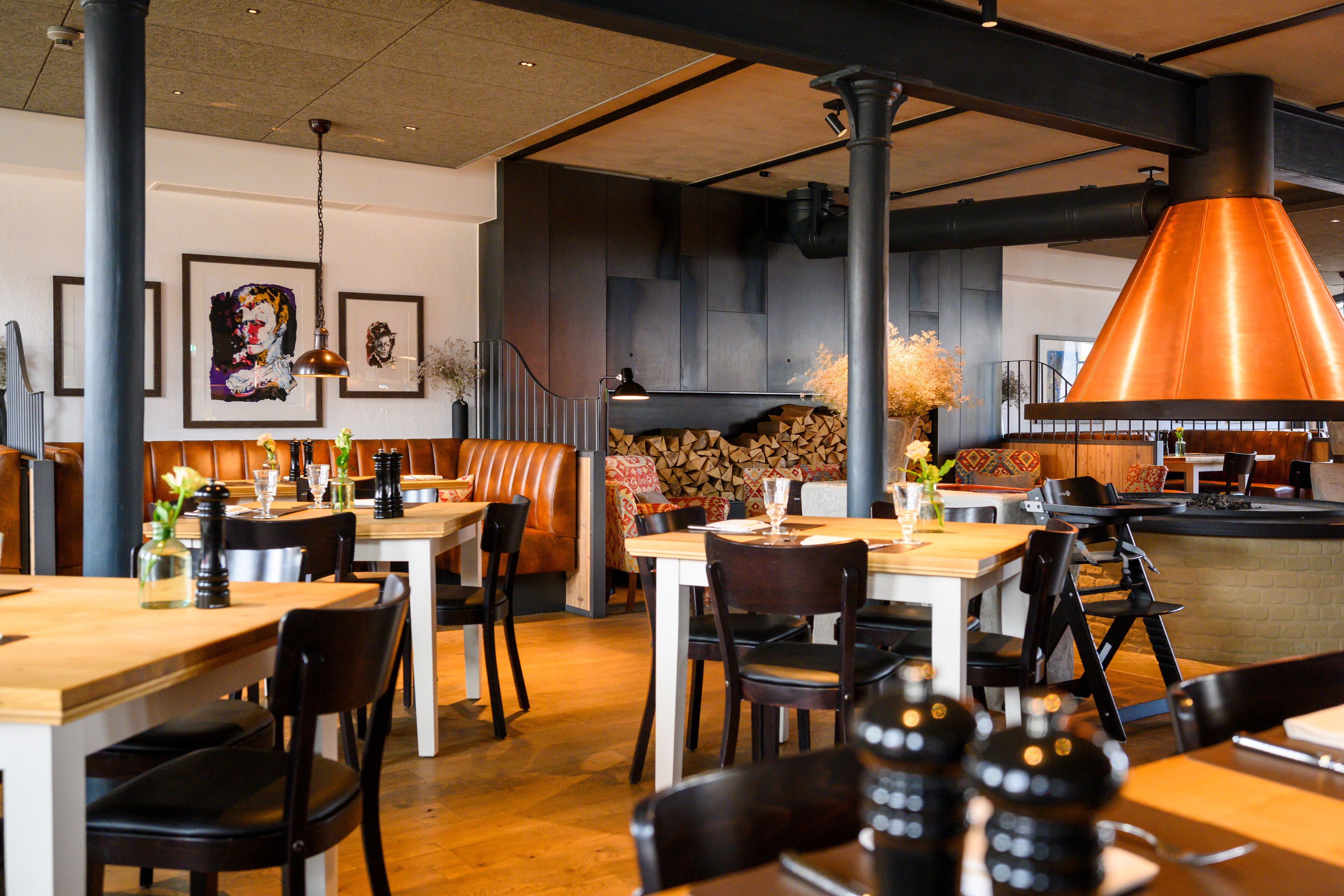 L’intérieur d’un restaurant avec des tables en bois, des chaises noires et un grand luminaire de style industriel, de couleur cuivrée.