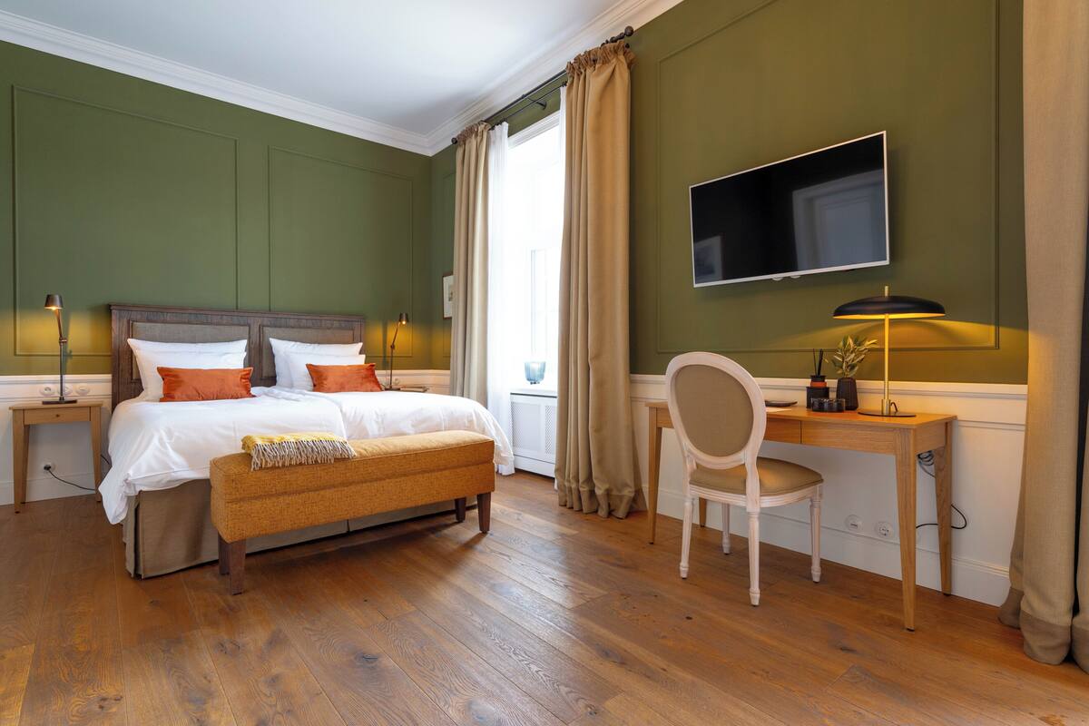 A bedroom with a wooden bed, a desk, a chair, a TV, and a window with curtains.