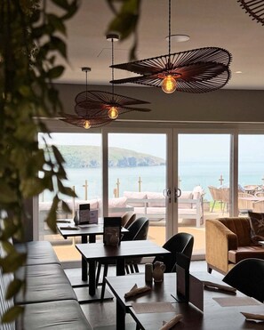 A modern dining area with a view of the sea, featuring pendant lights, a table with a menu, and comfortable seating. - The Gwbert Hotel (Cardigan)
