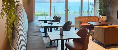 A modern dining area with a view of the sea, featuring a long bench, tables, and hanging light fixtures.