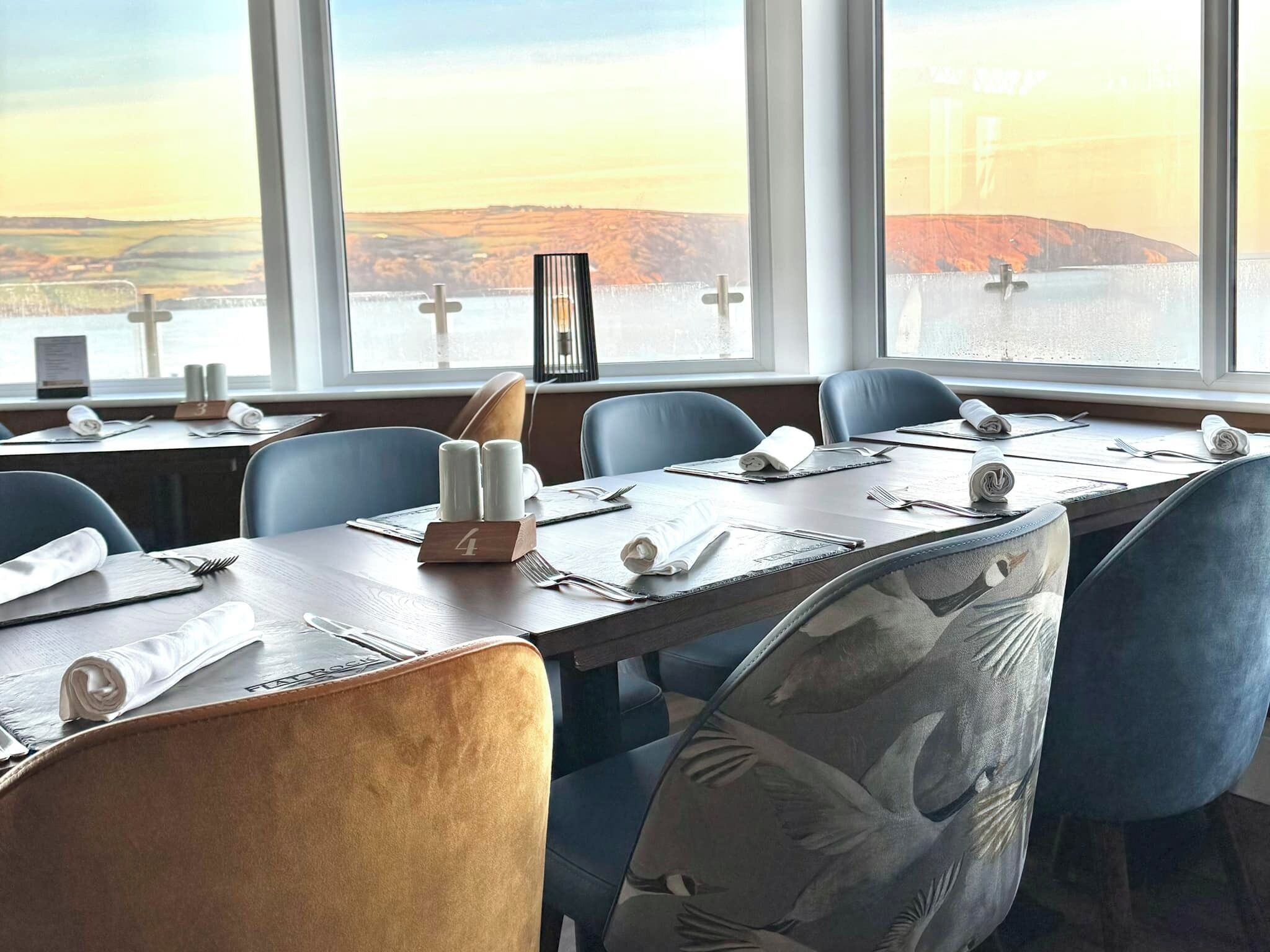 A dining area with tables set for four, chairs with floral upholstery, and a view of a landscape through large windows.