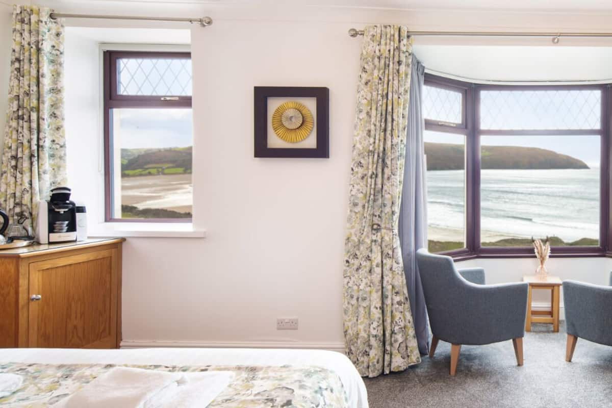 Ein Schlafzimmer mit einem Bett, einem kleinen Tisch mit Lampe, einem Sessel und Blick auf das Meer durch große Fenster.