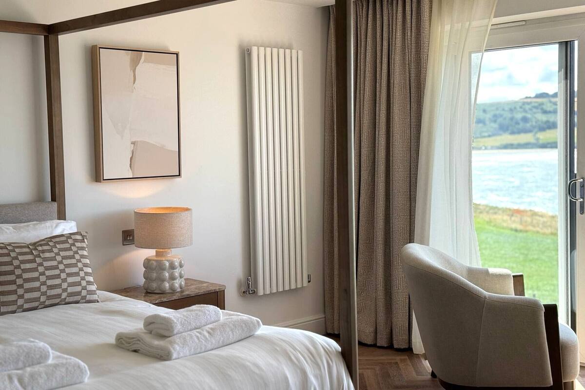 Ein Schlafzimmer mit einem Himmelbett, einem Nachttisch mit Lampe, einem Heizkörper und Blick ins Freie durch ein Fenster.