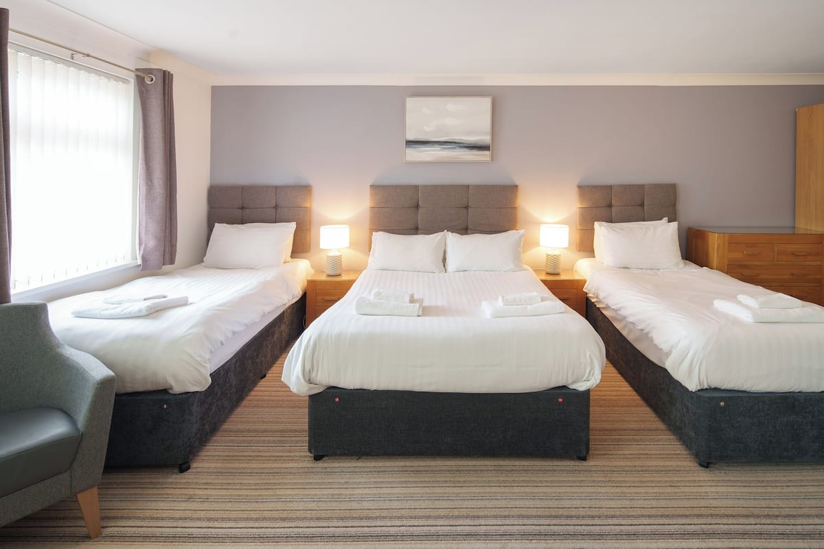 Ein Hotelzimmer mit drei Betten, einem grauen Sessel und einem Fenster mit Vorhängen.