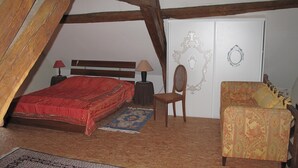 A bedroom with a wooden ceiling, a bed with a red bedspread, a chair, a small table with a lamp, and a patterned sofa. - Small rural  and charming house in a quiet hamlet. (Hérisson)