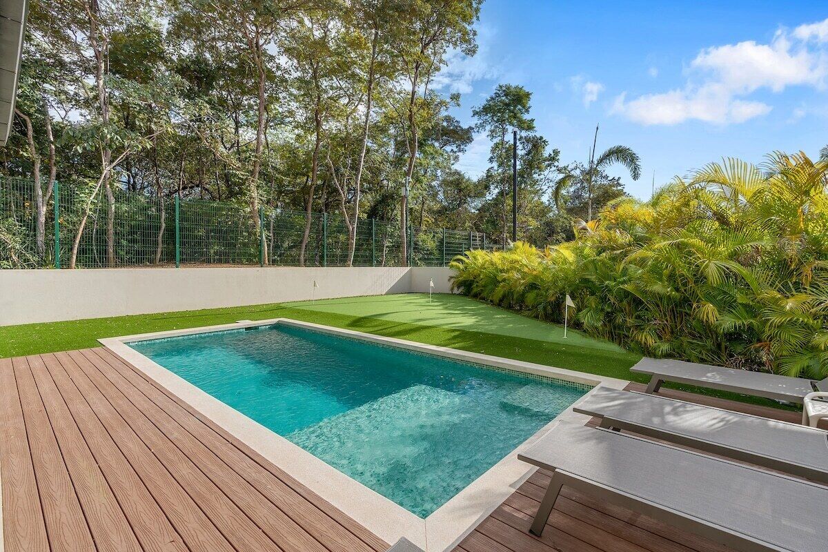 A rectangular swimming pool with a wooden deck, surrounded by a green fence and lush tropical vegetation.