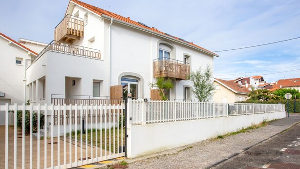 A two-story residential building with a white exterior, a red-tiled roof, and a white fence.