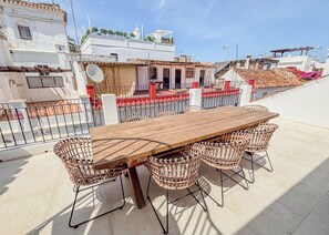 An outdoor patio with a wooden table and wicker chairs, surrounded by a metal fence, overlooking a residential area with white buildings and red-tiled roofs.