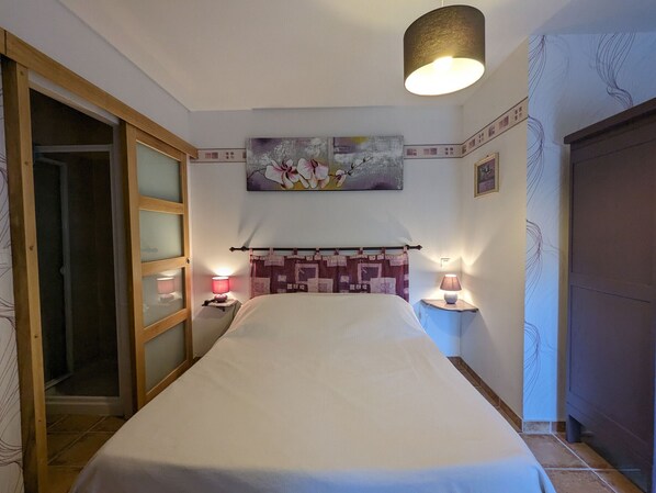 A bedroom with a bed, two bedside lamps, a painting on the wall, and a door with a glass panel. - Countryside Retreat with Spacious Grounds, Pool Access, and Family-Friendly Amenities (Pérassay)