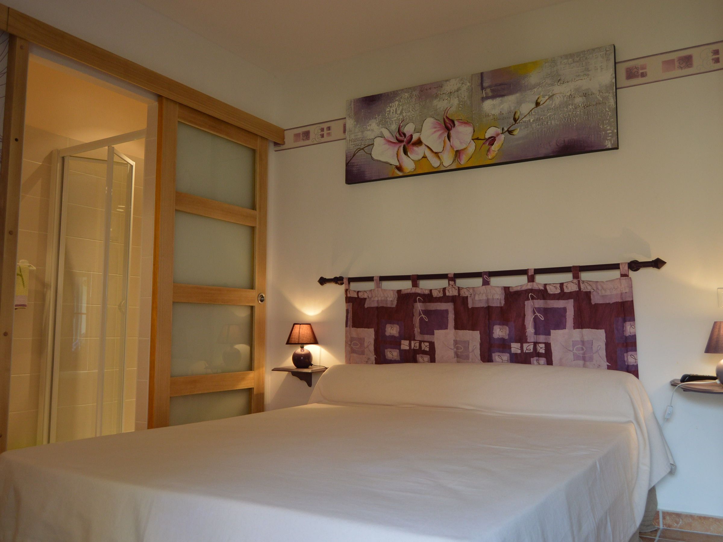 Une chambre à coucher comprenant un lit, une table de chevet, une lampe fixée au mur, une tête de lit matelassée et une porte-fenêtre coulissante.