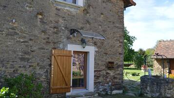 A stone building with a wooden door and window shutters, a small garden area with a stone wall and a metal gate, and a clear sky.