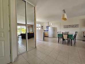 Un amplio salón con un ventanal corredera que da a un balcón, un comedor con mesa y sillas, y una cocina con refrigerador y salpicadero de azulejos.