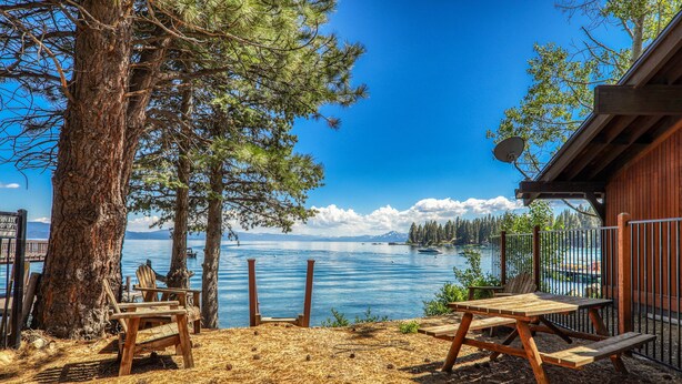 A wooden deck with chairs and a table overlooking a lake, surrounded by trees and a cabin. - Reflections Lakeview By Tahoe Getaways - Hot Tub & Buoy, Dog OK (Carnelian Bay)