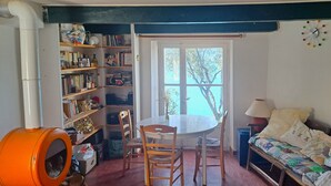 A cozy room with a wooden dining table, chairs, a bookshelf, a sofa, and a window with a view of trees. - "les Terrasses" : Maison Dans le Village, 4 Terrasses (Sainte-Croix-du-Verdon)