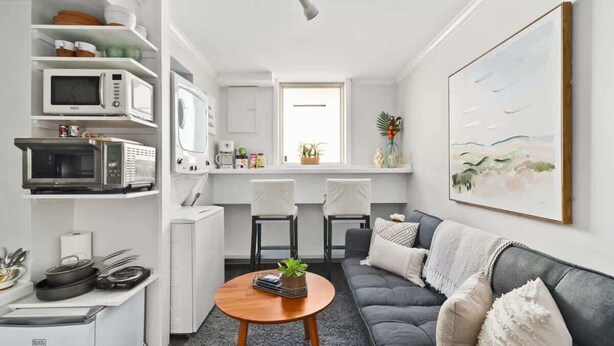 Un espace de vie compact comprenant un coin cuisine, un canapé gris, une table basse et un tableau accroché au mur.