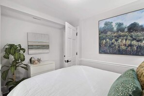 Ein Schlafzimmer mit einem großen Bett, einer weißen Kommode, einer Topfpflanze und einem Bild von einem Feld.