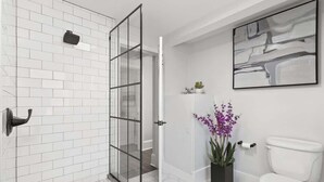 Une salle de bain avec des carreaux de céramique blancs rectangulaires, une douche en verre, des toilettes blanches et un pot de fleurs noir orné de fleurs violettes.