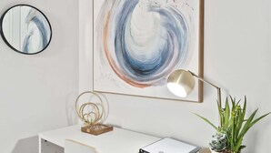 Un bureau moderne, agrémenté d’un grand tableau abstrait, d’une lampe de bureau, d’une plante en pot et d’un miroir.