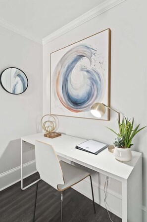 A modern office desk with a large abstract painting, a desk lamp, a potted plant, and a mirror.