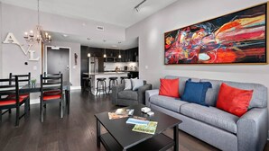 A modern living room with a grey sofa, red and blue cushions, a dining table with chairs, a coffee table with magazines, and a large abstract painting on the wall. - 2BR Condo w/ Balcony + Free Tix | Walk to Downtown (Asheville)