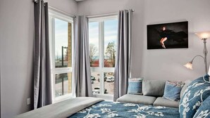 A bedroom with a bed, grey curtains, a grey sofa, a blue patterned bedspread, a wall-mounted painting, and a floor lamp. - 2BR Condo w/ Balcony + Free Tix | Walk to Downtown (Asheville)