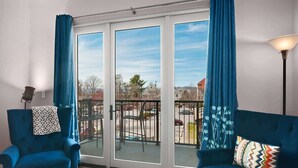 A room with a blue armchair, a sofa, and a balcony with a view of a parking lot and buildings. - 2BR Condo w/ Balcony + Free Tix | Walk to Downtown (Asheville)