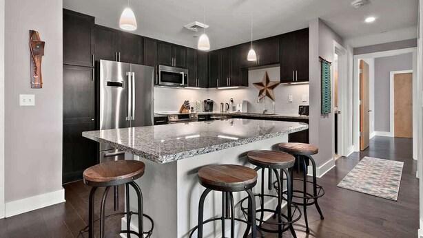 A modern kitchen with dark cabinets, stainless steel appliances, and a large island with bar stools. - 2BR Condo w/ Balcony + Free Tix | Walk to Downtown (Asheville)