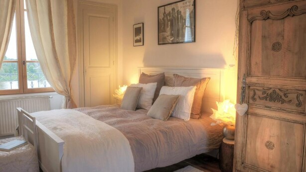 Um quarto com cama, mesas de cabeceira, cômoda, janela com cortinas e um quadro na parede.