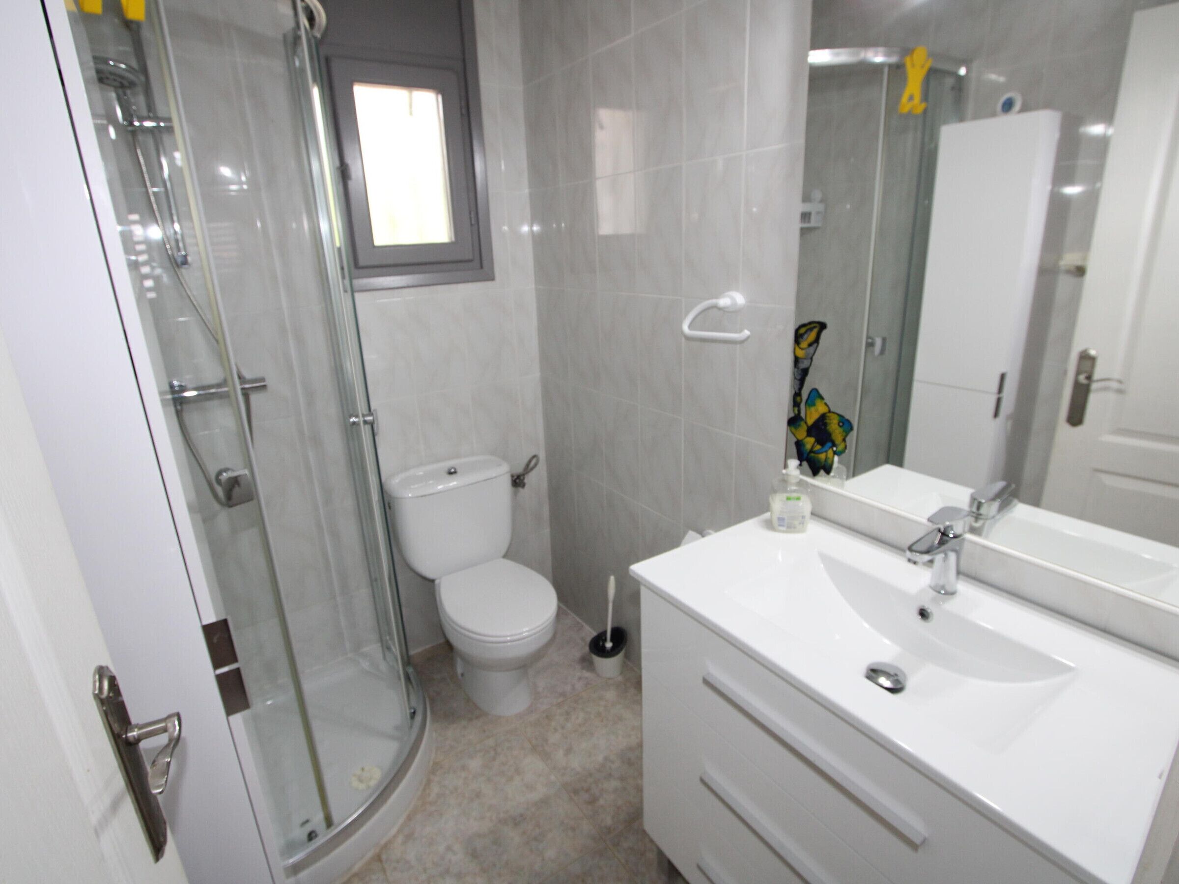 Ein Badezimmer mit Duschkabine aus Glas, Toilette, Waschbecken mit Spiegel und einem Fenster über der Toilette.