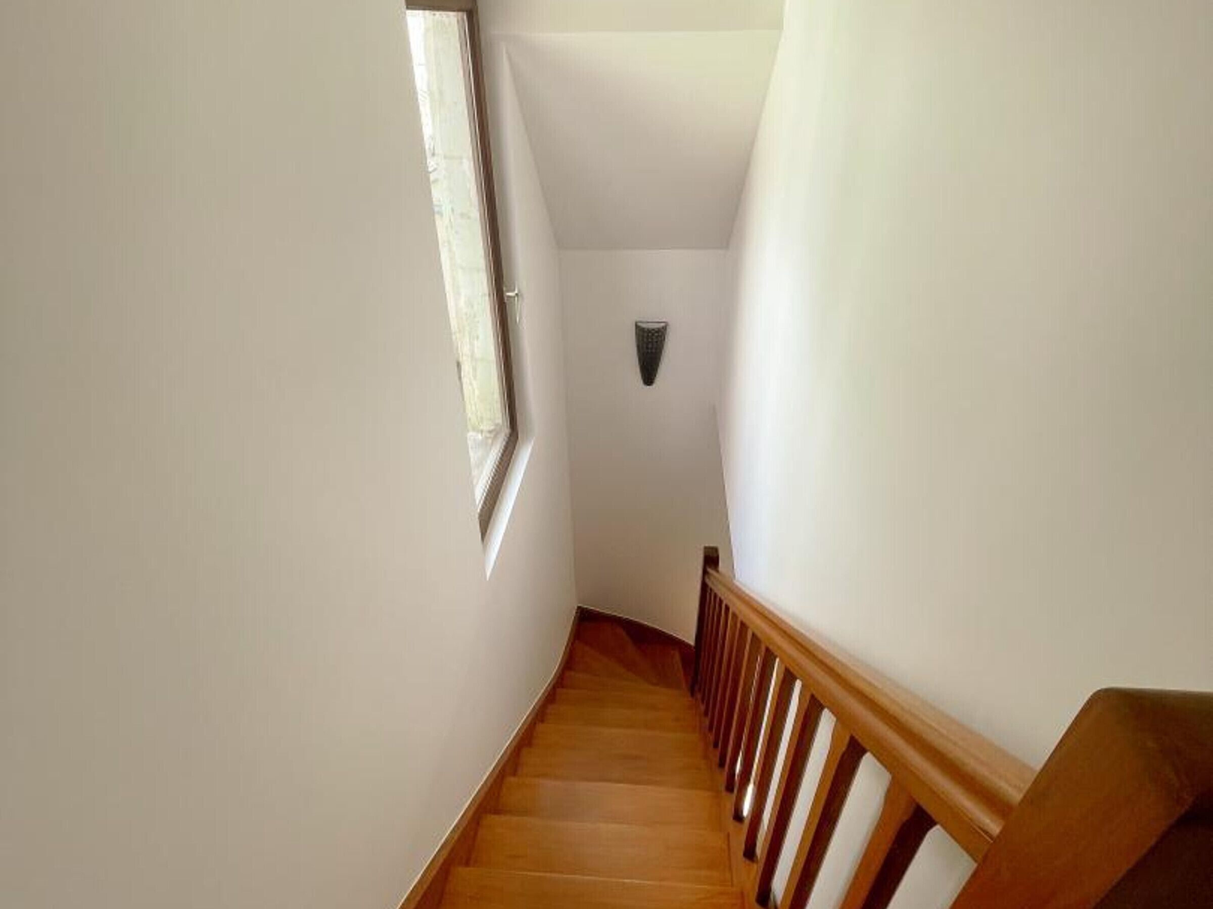 Un escalier étroit avec des marches en bois et une rampe en bois, menant à un palier où est fixée une petite applique murale.