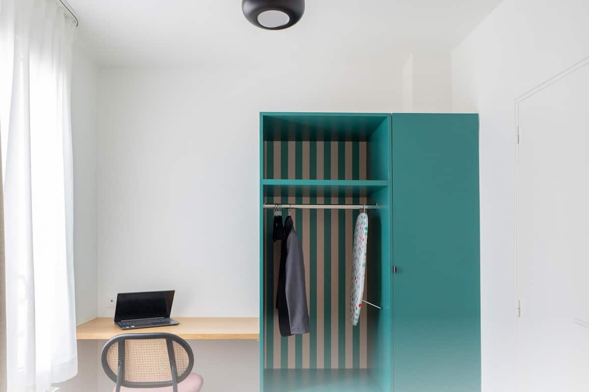 A room with a desk, laptop, teal wardrobe, and a chair.