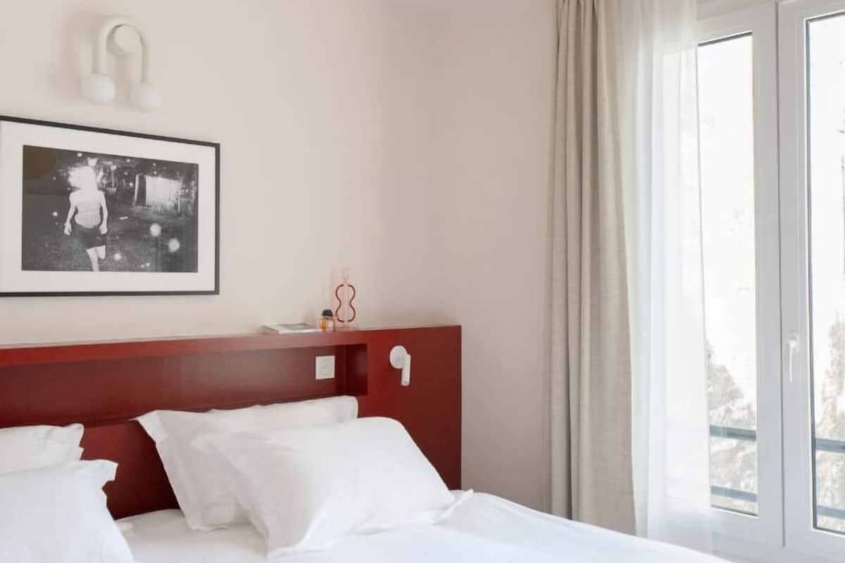 A hotel room with a bed, a framed picture on the wall, a ceiling light fixture, and a window with curtains.