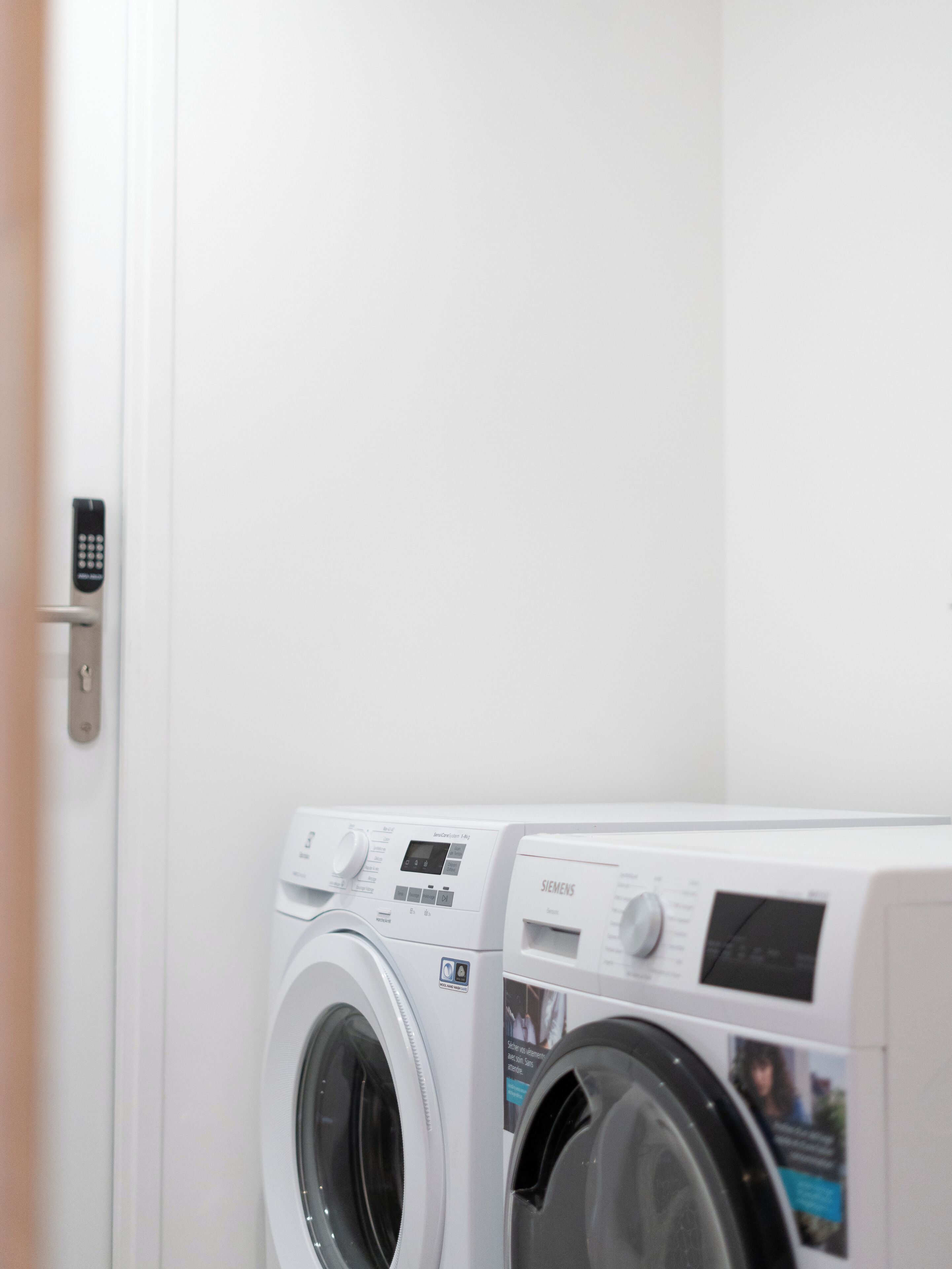 A front-loading washing machine in a laundry room with a door and a remote control on the wall.