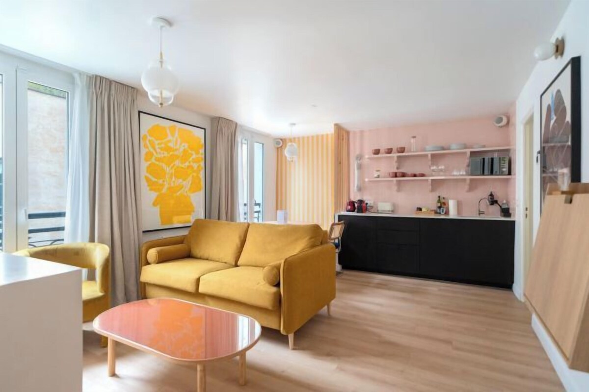 A modern living room with a yellow sofa, a wooden coffee table, and a kitchen area with a black countertop and shelves.