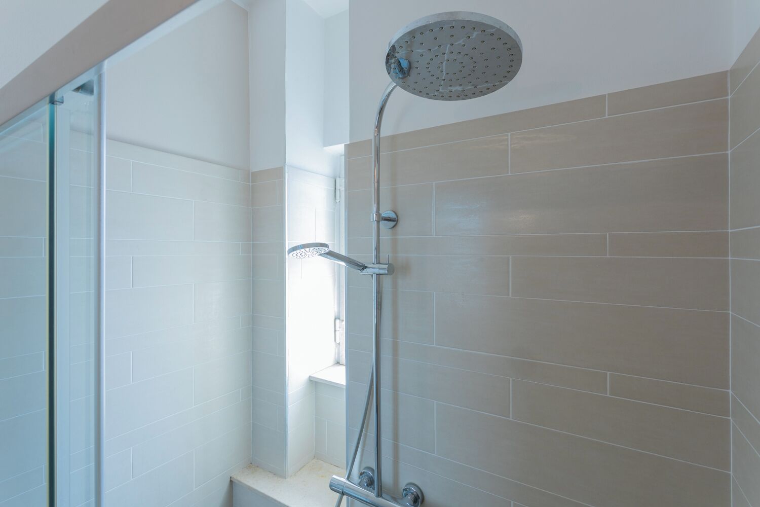 Ducha moderna con cabezal de ducha de lluvia y cabezal de ducha manual, paredes de azulejos blancos y puerta de cristal.