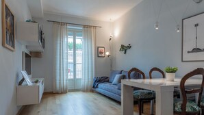A modern living room with a sofa, a dining table, and chairs. There is a window with curtains, a wall-mounted shelf with a plant, and a framed picture. - La Casa dei Viaggiatori by Wonderful Italy (Turin)