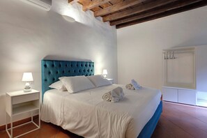 Ein ordentlich bezogenes Bett mit weißer Bettwäsche und einem blauen Kopfteil, ein Nachttisch mit Lampe sowie ein Kleiderschrank mit Spiegel.