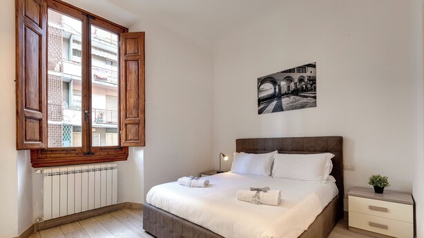 Une chambre avec un grand lit, une fenêtre avec des volets en bois, un radiateur, une table de chevet avec une lampe et un tableau encadré au mur.